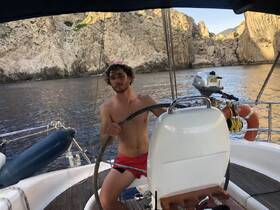 A young man navigates a sailboat, showcasing the beautiful rocky coastline in the background — a perfect moment of adventure on the water.