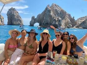 A joyful group of friends enjoys a sunny day on the water, celebrating with cocktails against the stunning backdrop of Cabo's iconic rock formations.
