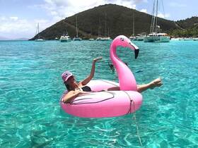 A woman enjoys a sunny day floating on a pink flamingo in crystal-clear waters, surrounded by boats — a perfect scene for summer fun and relaxation.