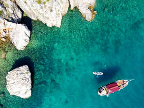 An aerial view captures a beautiful boat anchored in crystal-clear turquoise waters, with people enjoying the surroundings — a perfect scene for a summer getaway.