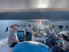 A lively group enjoys a fun day on the water, sharing food and drinks aboard a boat, with colorful inflatables and friends in the background.