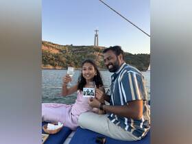 A couple celebrates on a sailboat, toasting with glasses of wine while showcasing a photo against a stunning backdrop of a landmark — perfect for romantic getaways or special occasions.
