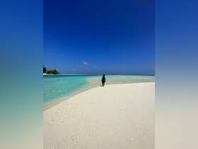 A lone figure walks along a pristine beach, surrounded by crystal-clear waters and a vibrant blue sky — a perfect snapshot of tropical paradise.