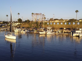 A picturesque view of the Oceanside harbor at sunset, featuring a sailboat gliding through calm waters, surrounded by a vibrant marina.
