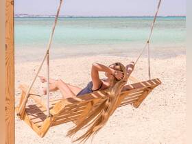 A woman relaxes on a wooden swing at the beach, enjoying the serene waters and sunshine — a perfect spot for unwinding and soaking up the sun.
