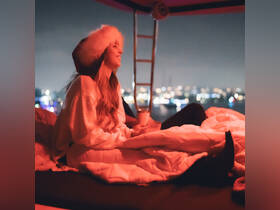 A woman enjoys a cozy evening on a boat, wrapped in blankets and sipping a drink, with a beautiful nighttime cityscape in the background.