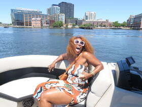 A joyful woman enjoys a sunny day on a boat, celebrating with a drink and laughter against a vibrant waterfront backdrop.