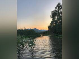 A serene sunset view over a tranquil waterway, framed by lush greenery and distant mountains — a perfect setting for peaceful boat rides and nature exploration.
