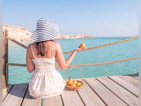 A woman enjoys a serene moment by the turquoise waters, holding an orange while seated on a wooden dock — a perfect snapshot of relaxation and natural beauty.