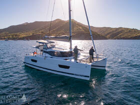 A catamaran sails through clear waters with two people on deck, showcasing the beauty of a day on the water amidst stunning coastal scenery.