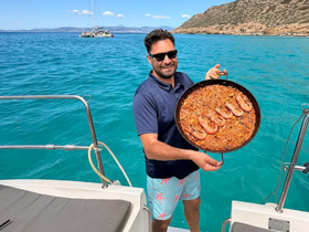 A chef proudly presents a delicious paella on the deck of a boat, showcasing the culinary delights available during a sailing adventure.