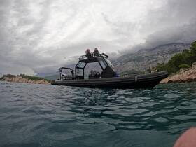 Two people enjoy a scenic ride on a sleek boat along a picturesque coastline, framed by dramatic mountains and cloudy skies.