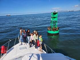 A lively group enjoys a day on the water, posing on the bow of a boat with sea lions lounging on a nearby buoy — a perfect snapshot of marine adventure.