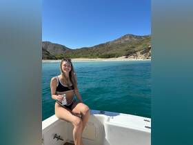 A woman enjoys a refreshing drink while relaxing on the edge of a boat, with stunning beach scenery in the background — perfect for a day of fun on the water.