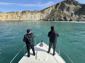 Two friends enjoy a fishing trip on a boat, casting their lines into the clear blue waters against a stunning coastal backdrop.