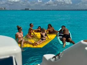 A lively group enjoys a sunny day on the water, lounging on a floating mat and paddleboarding in the vibrant turquoise sea — perfect for summer adventures.