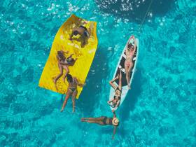 A lively group enjoys a sunny day on the water, relaxing on a floating mat and paddleboard in crystal-clear turquoise waters — perfect for summer fun and social gatherings.