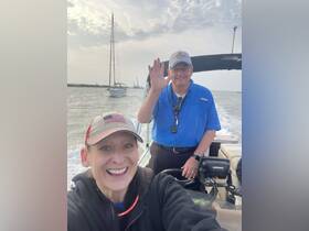 A cheerful selfie of two individuals aboard a boat, with a sailboat visible in the background — capturing the joy of a day on the water.