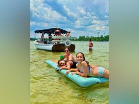 A joyful group of friends enjoys a sunny day on the water, lounging on a floating mat while a pontoon boat is anchored nearby — perfect for summer fun and relaxation.