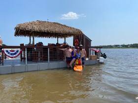 A lively group enjoys a fun day on the water, posing on a floating mat with a tiki hut backdrop — perfect for summer celebrations and family outings.