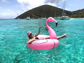 A woman enjoys a sunny day floating on a pink flamingo in crystal-clear waters, surrounded by stunning island scenery — perfect for a relaxing beach getaway.