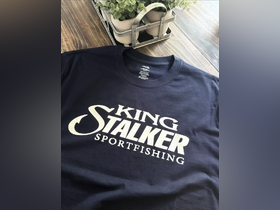 A close-up shot of a navy t-shirt featuring the logo 'King Stalker Sportfishing' on a wooden surface, surrounded by a plant. Ideal for fishing enthusiasts.