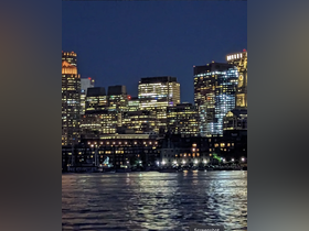 A nighttime cityscape featuring illuminated buildings reflecting on the water, creating a vibrant urban atmosphere.