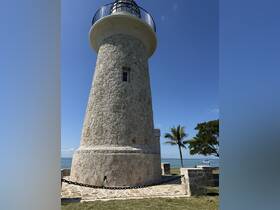 A lighthouse stands tall against a clear blue sky, surrounded by lush greenery and a serene waterfront, creating a picturesque coastal scene.