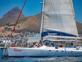 A lively group enjoys a sailing experience aboard the catamaran 'Tigresse' against a stunning backdrop of mountains and coastal scenery.