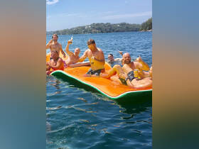 A lively group of friends enjoys a sunny day on a floating mat, celebrating with drinks in the water — perfect for summer fun and memorable outings.
