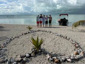 A group of friends poses on the beach with a boat in the background, surrounded by a decorative shell arrangement — a perfect moment capturing fun and friendship by the water.