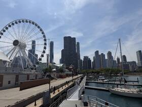 A view of a marina with a skyline backdrop, featuring a Ferris wheel and several boats docked. The scene captures the essence of urban waterfront life.