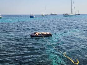 A person relaxes on a floating mat in crystal-clear waters, surrounded by boats — a perfect scene for a summer getaway or beach day.