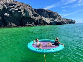 Two friends relax on a floating mat in vibrant green waters, enjoying a sunny day surrounded by stunning rocky cliffs — a perfect setting for a summer getaway.