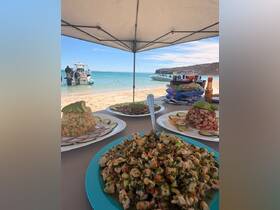 A delicious spread of seafood dishes is laid out on a beach, perfect for a relaxing day by the water. Guests enjoy fresh flavors while boats are anchored nearby.