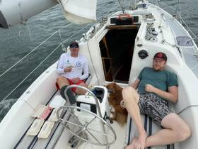 Two friends relax on a sailboat with a dog, enjoying snacks and drinks while sailing — a perfect day on the water.