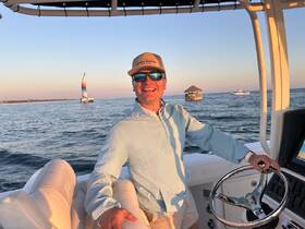 A cheerful captain navigates a boat during sunset, with a colorful sailboat in the background, capturing the essence of a perfect day on the water.
