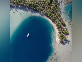A stunning aerial view of a yacht anchored in a serene blue lagoon, surrounded by lush palm trees and a pristine sandy beach — an idyllic setting for a tropical getaway.