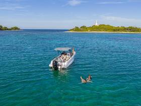 A group enjoys a refreshing swim in crystal-clear waters near a boat, with a picturesque island and lighthouse in the background — perfect for island hopping adventures.