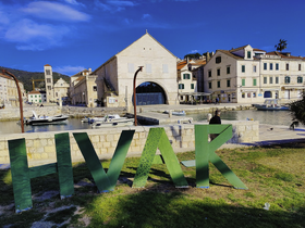 A scenic view of a harbor with buildings and boats, featuring large green letters spelling 'HVAK' in the foreground.