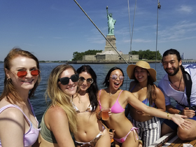 A lively group of friends enjoys a sunny day on the water, celebrating with drinks and the iconic Statue of Liberty in the background — perfect for summer outings in New York.