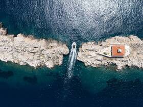 A stunning aerial view of a boat navigating near a rocky coastline with a lighthouse, showcasing the beauty of the Mediterranean landscape.