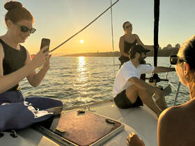 A group of friends enjoys a beautiful sunset sailing experience, capturing memories on their phones as they relax on the deck of a sailboat.
