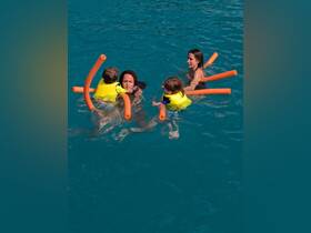 A joyful family enjoys swimming in clear turquoise waters, with children using pool noodles for support — a perfect moment of fun and bonding in the sun.