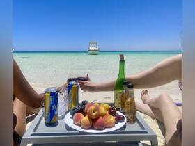 Friends enjoy a beachside celebration with drinks and fresh fruit, while a boat is anchored in the background — perfect for a relaxing day on the water.