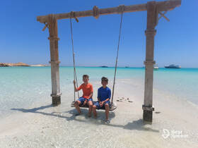 Two children enjoy a swing on a beautiful beach, with boats anchored in the turquoise waters — a perfect scene for family fun and relaxation.