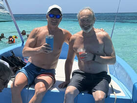 Two friends enjoy cocktails on a boat while swimming in crystal-clear waters, capturing the essence of a fun day on the water.