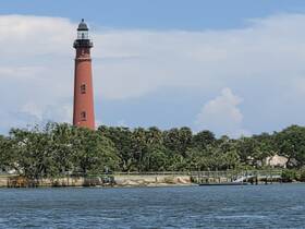 A striking lighthouse stands tall against a blue sky, surrounded by lush greenery, making it a notable landmark along the waterfront.