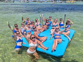 A lively group of friends enjoys a fun day on a floating mat in clear waters, celebrating with drinks and laughter — perfect for summer parties and memorable outings.
