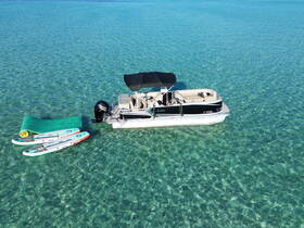A pontoon boat anchored in clear waters, accompanied by paddleboards and a floating mat, perfect for a day of fun on the water.